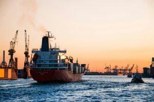 Red cargo ship Charles Island leaving an industrial port at sunset, cranes on shore and a tugboat nearby