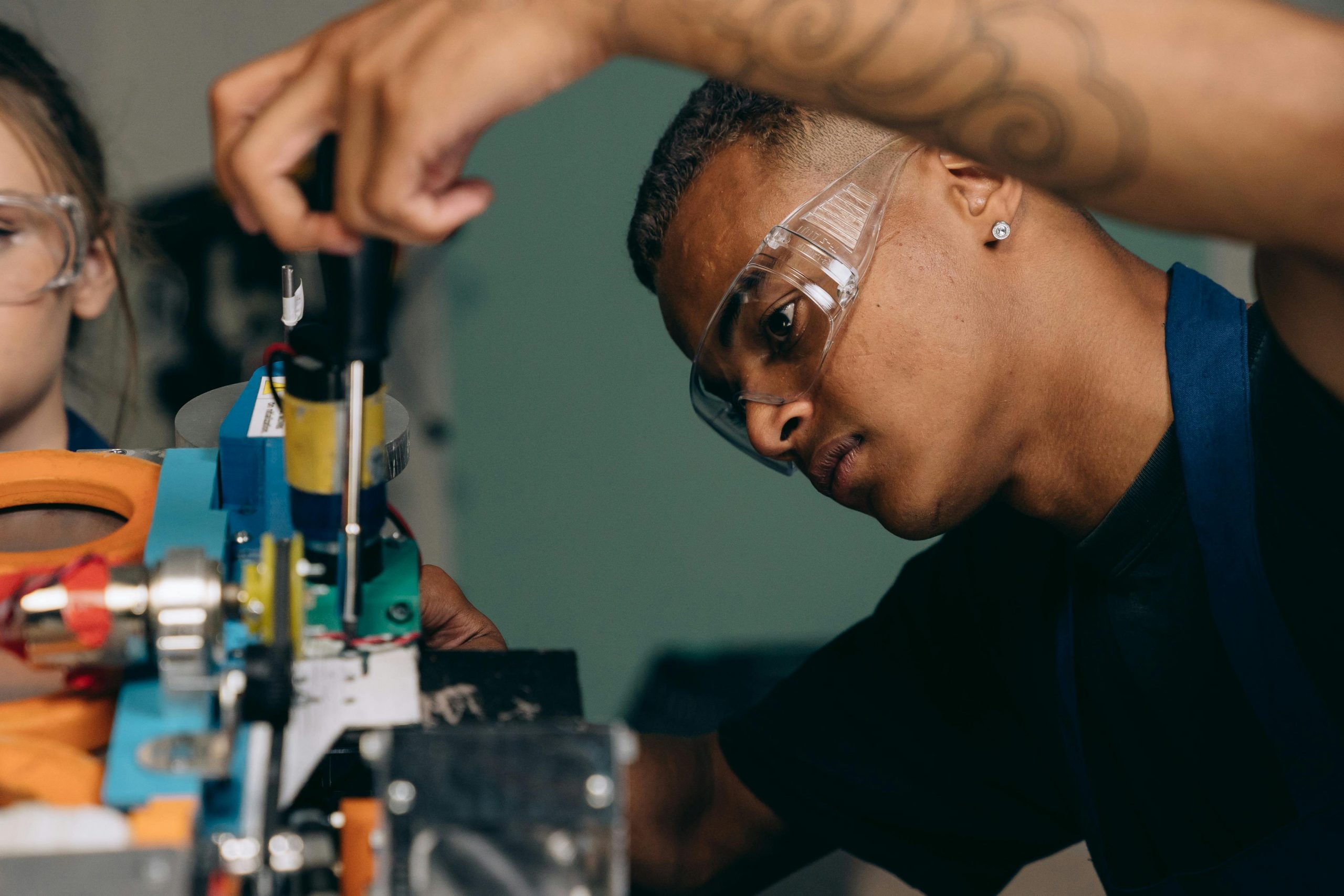 Young man wearing safety goggles carefully uses a screwdriver to adjust components on a blue machine.