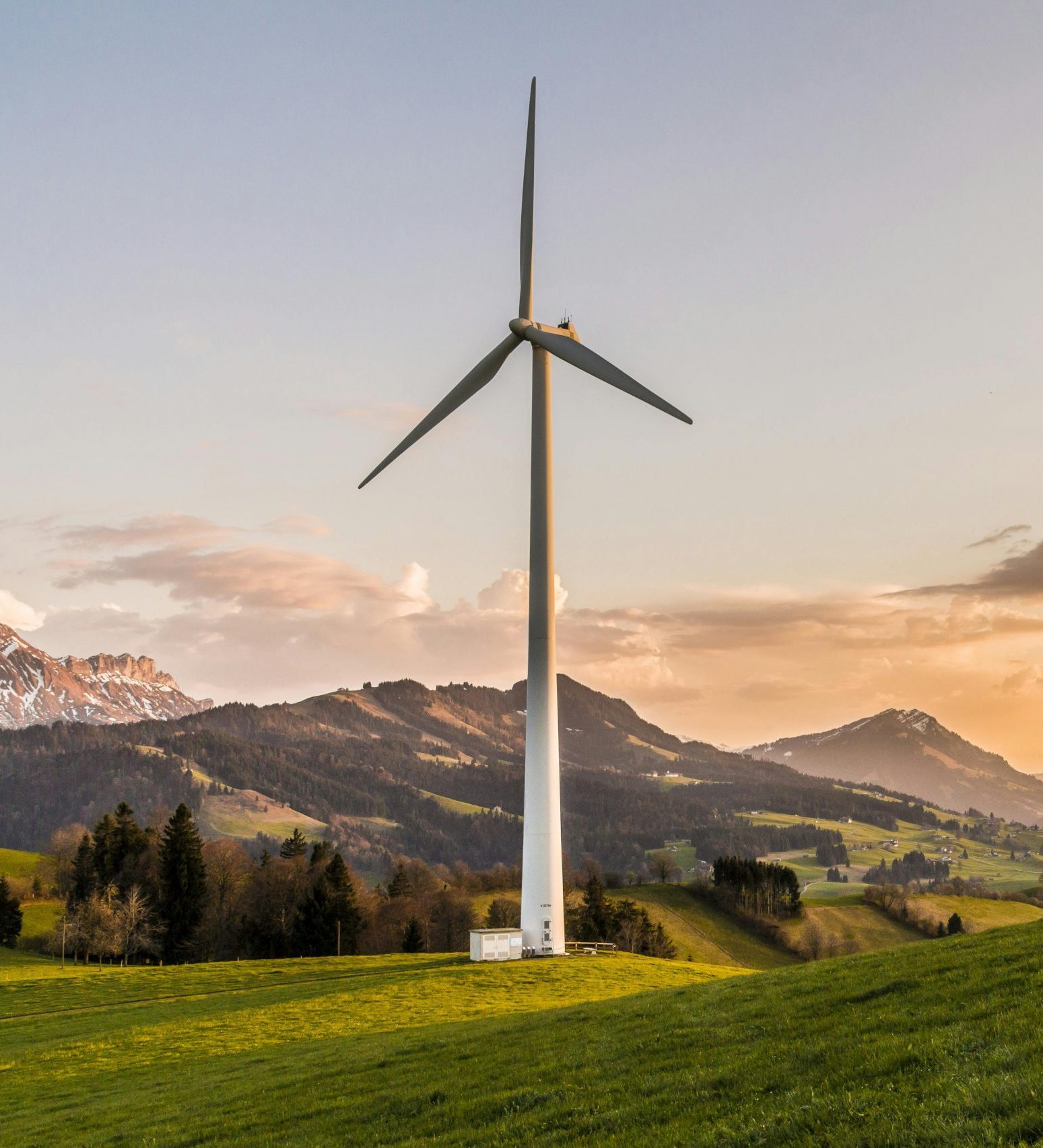 White three-blade wind turbine on a green hill beside a small shed, with rolling hills and snow-capped mountains at sunset.