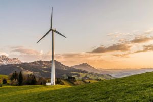 Single tall wind turbine on a green hillside overlooking rolling fields and distant mountains at golden sunset.