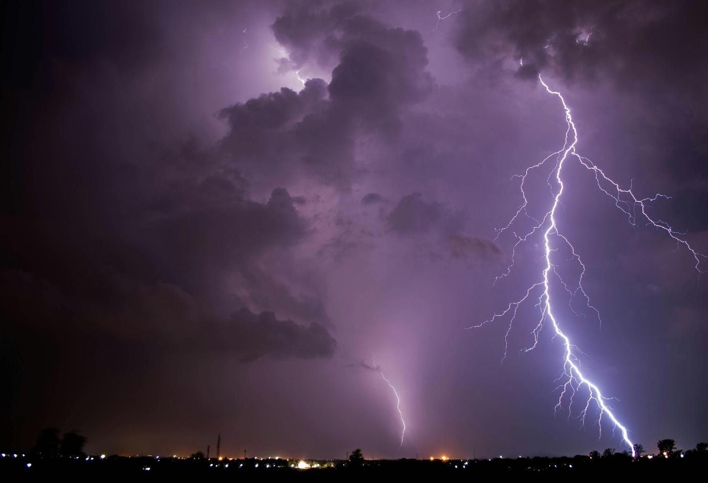 Forked lightning cutting through purple storm clouds above distant city lights at night.