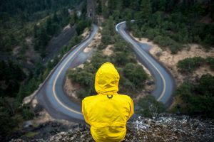 Person in a yellow rain jacket on a cliff edge looking down at a winding two-lane road through a forest.