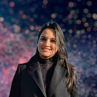 Smiling woman with long dark hair wearing a black coat and turtleneck, blurred colorful bokeh lights in the background.