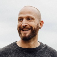 Smiling bald man with a trimmed beard and small earring wearing a dark crewneck sweater against a pale neutral background