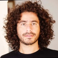 Head-and-shoulders portrait of a man with shoulder-length curly dark hair, light beard, neutral expression, and a plain light background.