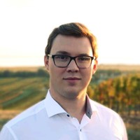Young man with short dark hair and glasses wearing a white shirt, standing outdoors with fields and a sunset in the background