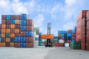 Stacks of colorful shipping containers at a busy port yard with a container handler lifting an orange container under a blue sky
